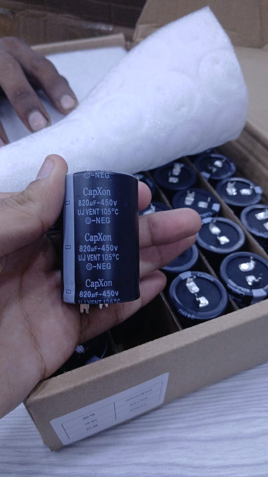 Hand holding CapXon electrolytic capacitor 820uF 450V near box of identical power supply capacitors
